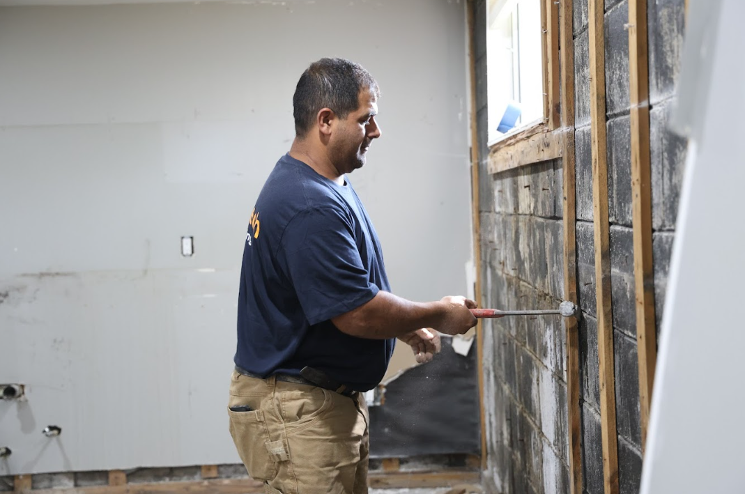 Aquatech Waterproofing technician in a branded dark shirt working on a mold-damaged basement wall near a window