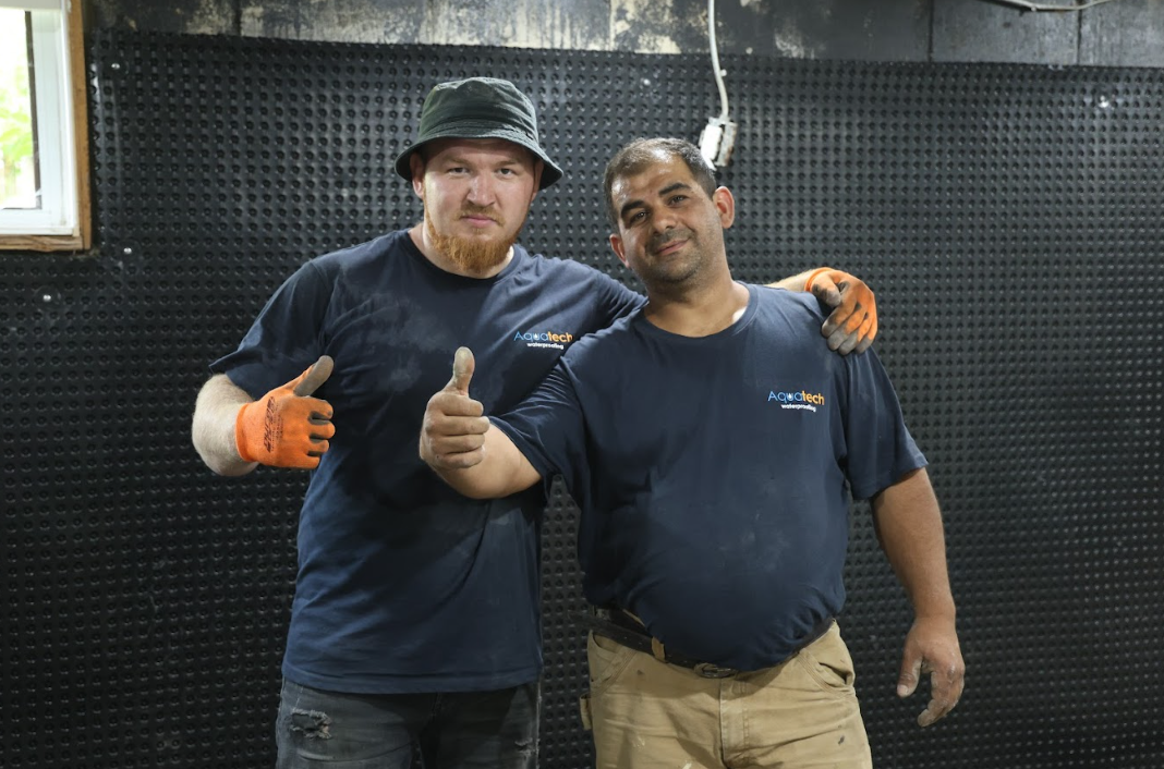 Aquatech Waterproofing technicians giving a thumbs-up in a basement with a black dimpled waterproofing membrane wall and a window