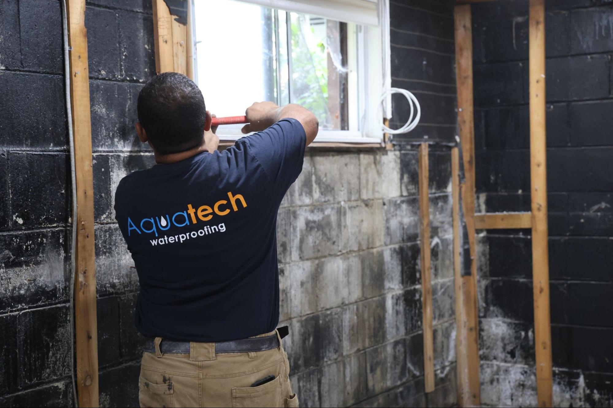 Aquatech Waterproofing technician scraping a basement concrete block wall near a window